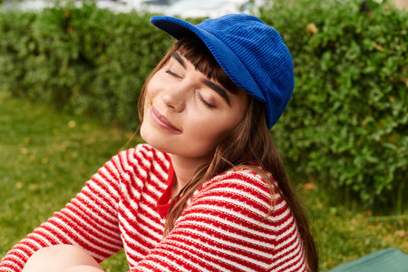 Young woman in a red striped sweater and blue cap relaxes in the sun, lost in thought and bliss.の写真素材