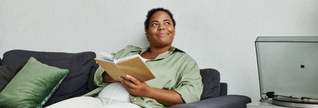 An African American woman enjoys her time at home, deeply engaged in reading a book on a couch, bannerの写真素材