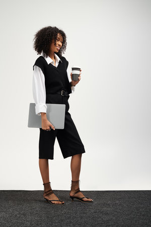 Charming young woman in business attire holds a laptop and coffee cup, smiling.の写真素材