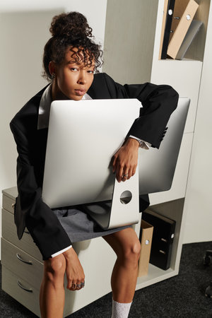 A stylish young woman confidently sits in a chic office, holding a sleek monitor.の写真素材