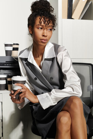 A confident and elegant young woman sits in a stylish office, sipping coffee while deep in thought.の写真素材