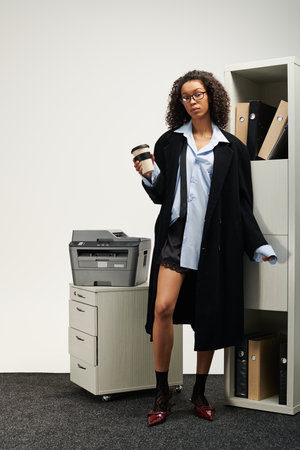 Young woman with curly hair holds a coffee cup while posing confidently in a stylish office setting.の写真素材