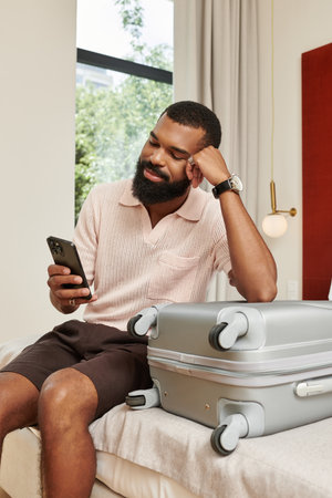 A stylish Black man relaxes in a chic hotel room, checking his phone with a suitcase nearby.の写真素材