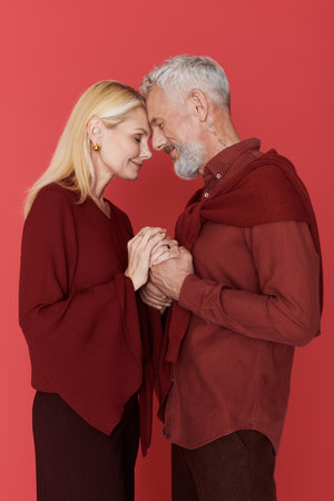 A senior couple in stylish maroon outfits shares a loving gaze against a vibrant backdrop.