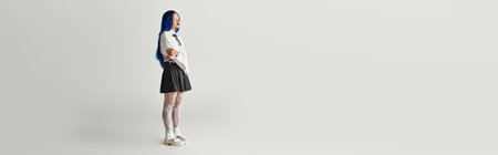 A young Asian woman with vibrant blue hair showcases her fashionable outfit against a grey backdrop,...
