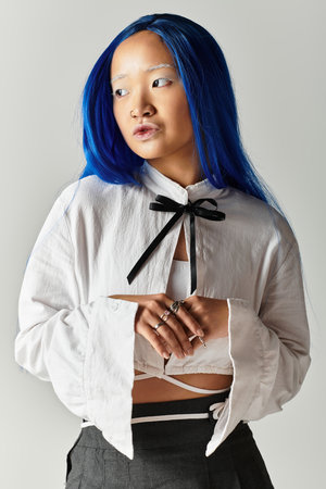 A young Asian woman with blue hair poses stylishly in trendy attire against a grey backdrop.