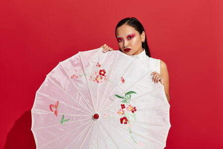 Young asian woman with striking eye makeup poses confidently against a bold red background.の写真素材