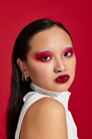 A young Asian woman showcases bold eye makeup and trendy attire in a striking red backdrop.