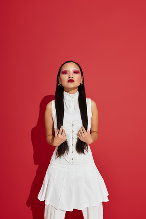 A confident young Asian woman poses in a red studio, highlighting her stylish outfit and eye makeup.の写真素材