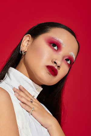A young Asian woman highlights her unique style and vivid eye makeup in a bold red background.の写真素材