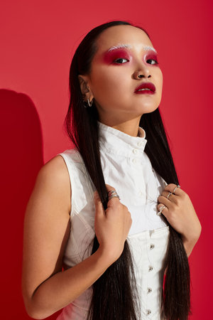 A young Asian woman showcases her stylish outfit and striking makeup against a red backdrop.の写真素材