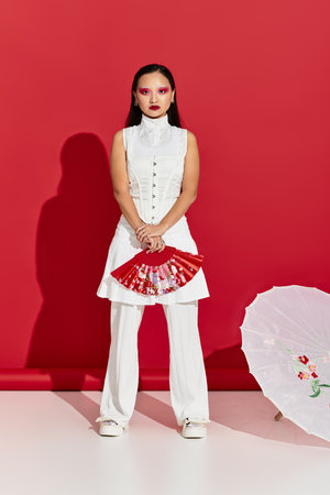 Young asian woman in trendy attire poses confidently with a fan in a vibrant red studio.
