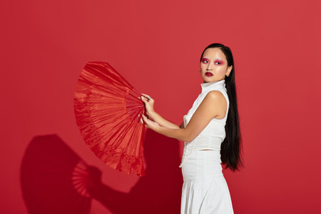 A young asian woman showcases her artistic eye makeup and fashionable outfit against a vivid red backdrop.の写真素材