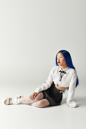 A stylish woman with blue hair poses on the floor of a grey studio, flaunting her outfit.の写真素材