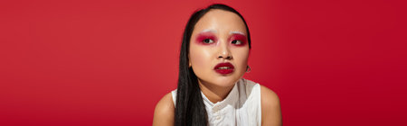 A young Asian woman exudes confidence with bold eye makeup, set against a striking red backdrop, bannerの写真素材