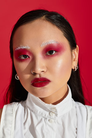 A young asian woman showcases her bold eye makeup and stylish outfit in a red studio.
