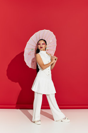 A young Asian woman showcases her trendy outfit and stunning eye makeup under a bright red backdrop.の写真素材