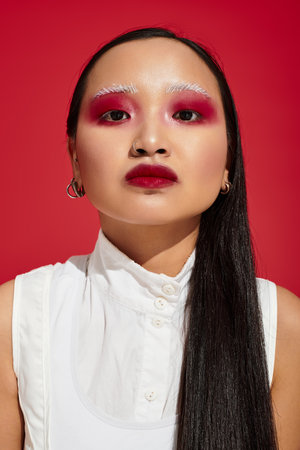 A young Asian woman showcases artistic eye makeup and a stylish outfit in a vivid red studio.の写真素材