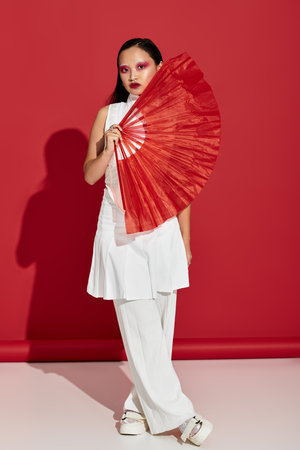 A beautiful young woman displays stylish fashion and bold eye makeup, holding a red fan.の写真素材