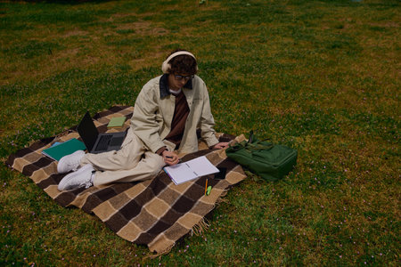 Young student relaxes on a blanket, writing notes while listening to music in a green park.