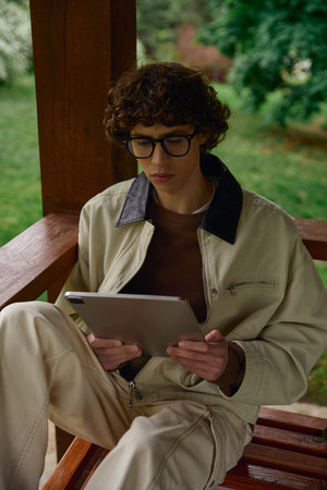 A young man dressed casually sits in a serene outdoor setting, focused on his tablet.