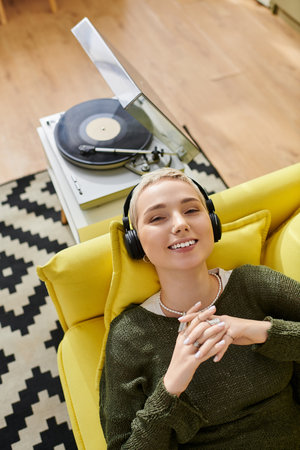 Young woman lies on a yellow sofa, smiling while listening to vinyl records at home.