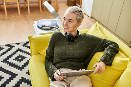 A young woman smiles on a yellow couch, relaxing and engaged with her tablet.の写真素材