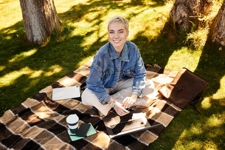 A young woman with short hair sits on a blanket in a green park, using her laptop.