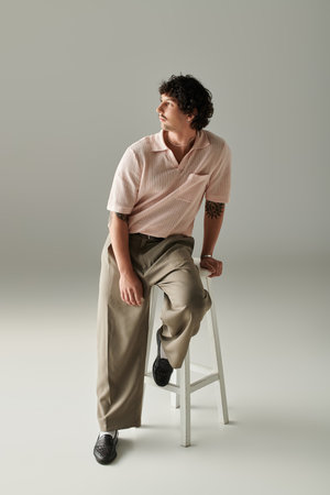 A stylish young man sits on a stool in a neutral toned space, dressed in trendy fashion attire.