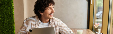 In a cozy cafe, a young man smiles while focused on his laptop and savoring a delightful drink.