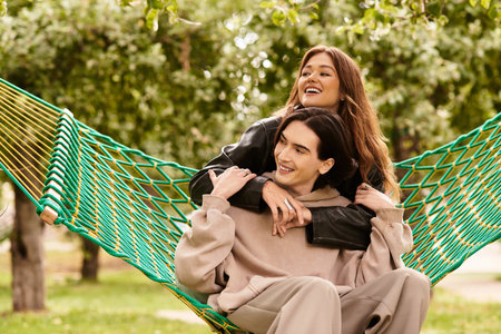 A loving couple shares a playful moment, laughing joyfully in a hammock surrounded by trees.
