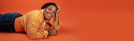 A fashionable plus size woman poses on an orange backdrop, flaunting her trendy autumn attire,...