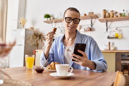 A young handsome man with feminine features smiles while eating breakfast and using his phone.