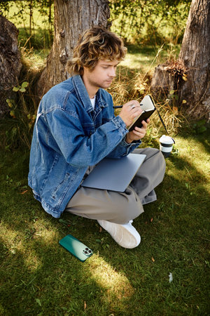 A handsome man in casual attire sits on the grass, writing in a notebook, surrounded by trees.