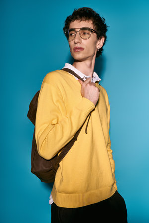 A fashionable young man stands confidently, wearing a yellow sweater and glasses, with a backpack.
