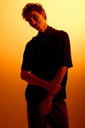 A handsome young man stands confidently in stylish attire, illuminated by a warm, glowing backdrop.