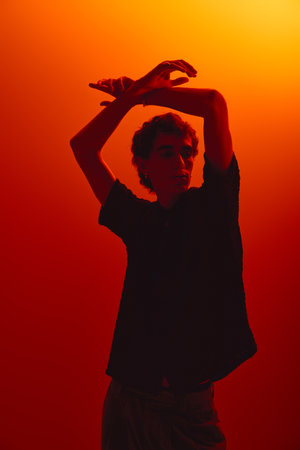 A handsome young man in stylish attire strikes a pose in a warm orange backdrop, exuding confidence.