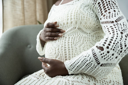 A pregnant African American woman gently cradles her belly while relaxing at home.