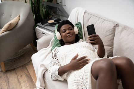 A pregnant African American woman enjoys music while relaxing at home on a cozy sofa.の写真素材