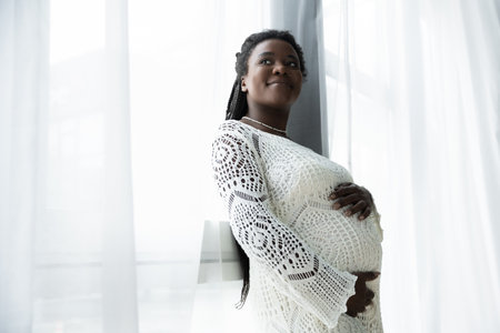 A pregnant African American woman with braided hair enjoys peaceful time at home, smiling gently.のeditorial素材