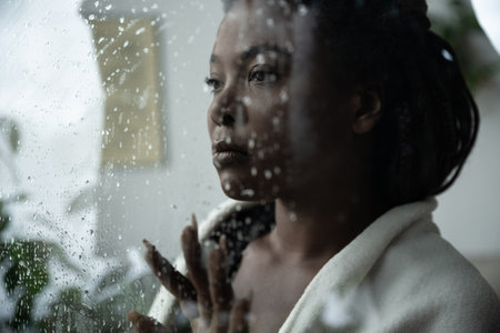 A woman with braided hair reflects deeply by the window, feeling overwhelmed by sadness at home.のeditorial素材