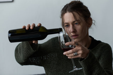 A young woman, feeling down, pours herself a glass of wine in her cozy living room at home.