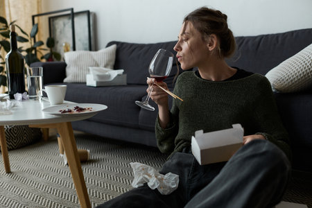 A young woman sits on the floor of her home, savoring wine and contemplating her thoughts.