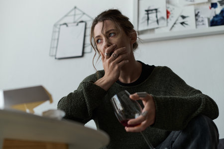 In a cozy living room, a young woman sits with a glass of wine, reflecting on her struggles.のeditorial素材
