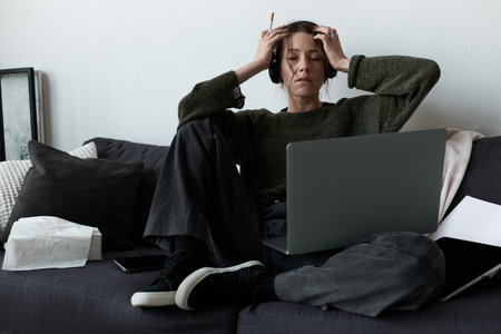 Woman experiences sadness and stress at home, seated on a couch with headphones on.