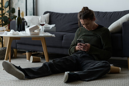 A young woman sits on the floor of her living room, absorbed in her phone while feeling upset.
