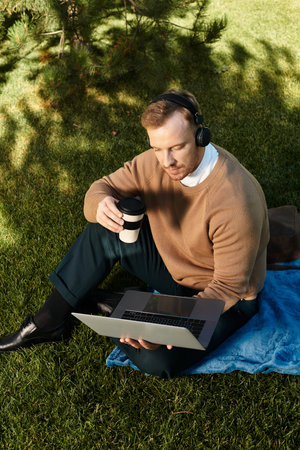 A young man sits on a blue blanket in a green area, working on his laptop and sipping coffee.