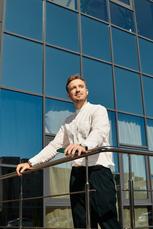 A young man poses by a railing, basking in the sunlight next to a glass building.のeditorial素材