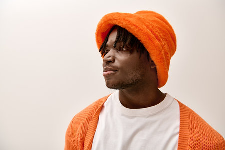 A stylish young man in a bright orange hat and knit cardigan stands confidently.