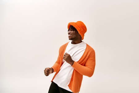 A young man wears an orange cardigan and hat while dancing with a cheerful smile indoors.